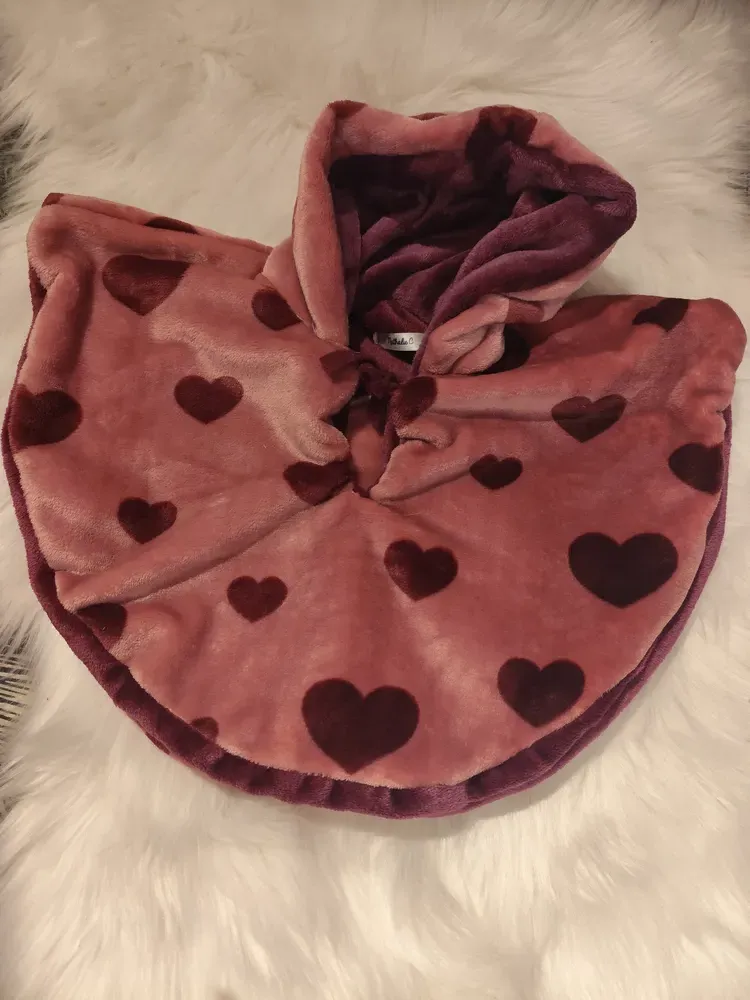 Cape polaire réversible pour bébé, face vieux rose ornée de cœurs violine, capuche relevée, posée sur une peluche blanche