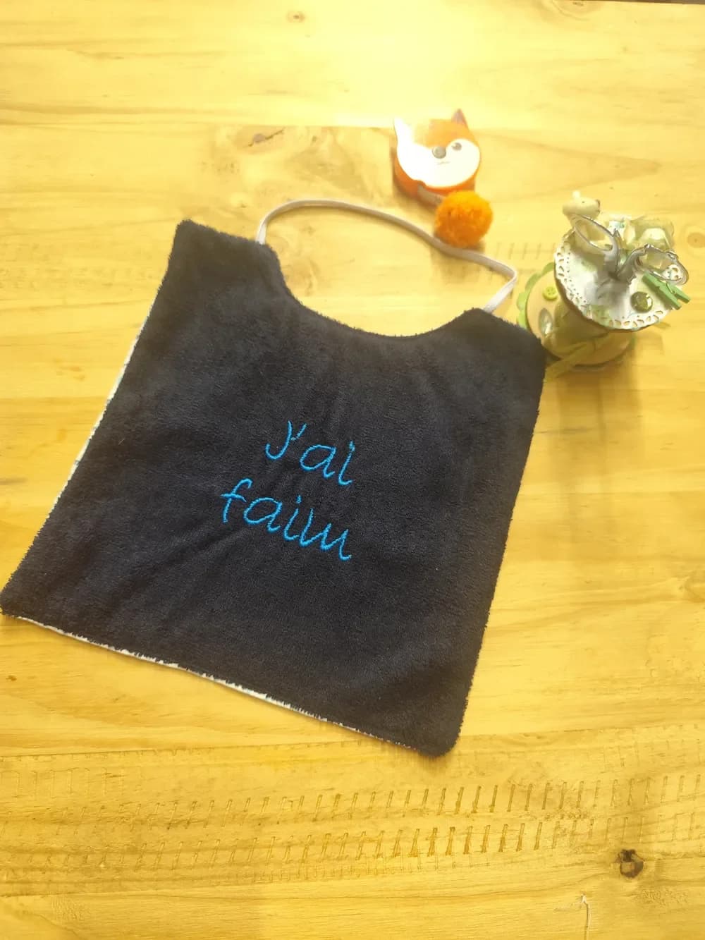 Bavoir en éponge bleu marine posé en biais sur une table en bois clair, brodé en fil bleu turquoise sur deux lignes des mots « J'ai faim » en lettres manuscrites, avec un élastique gris clair pour passer autour du cou. Derrière, un mètre ruban en forme de renard orange, un pompon orange et un pot à ciseaux décoré