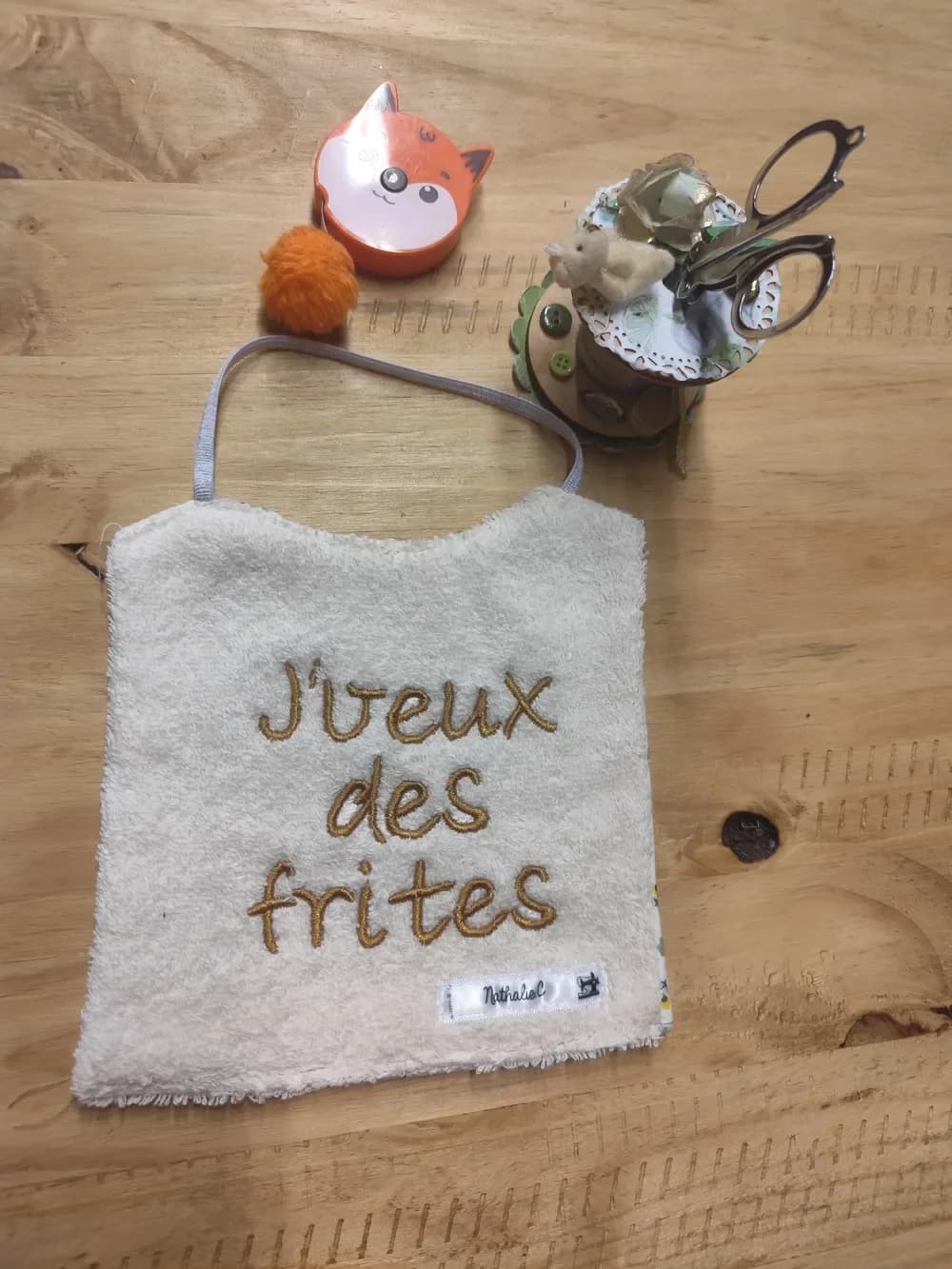 Bavoir en éponge écrue posé à plat sur une table en bois, brodé en fil doré sur trois lignes du message « J'veux des frites » en lettres manuscrites, avec une étiquette tissée « Nathalie C. » en bas à droite et un élastique gris clair pour passer autour du cou. Autour, un mètre ruban en forme de renard orange, un pompon orange et un pot à ciseaux décoré
