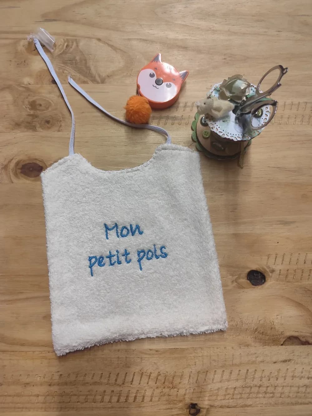 Bavoir en éponge écrue posé à plat sur une table en bois, brodé en fil bleu sur deux lignes des mots « Mon petit pois » en lettres arrondies, avec deux élastiques gris clair libres en haut, à nouer derrière la nuque. Autour, un mètre ruban en forme de renard orange, un pompon orange et un pot à ciseaux décoré