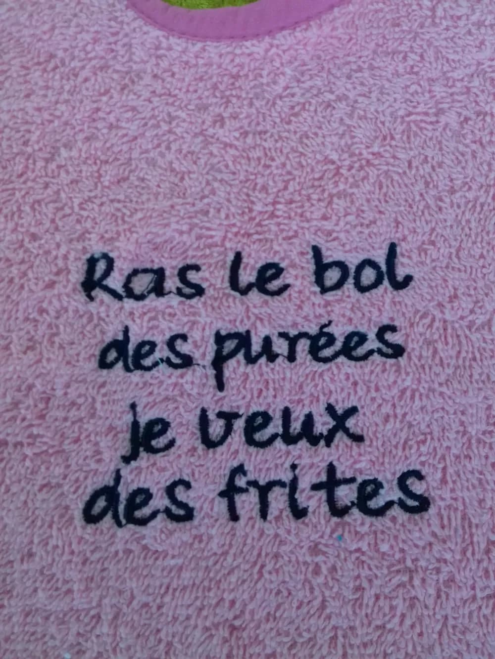 Gros plan d'un bavoir en tissu éponge rose bouclé, bordé d'un biais rose à l'encolure, brodé en fil bleu marine sur quatre lignes du message « Ras le bol des purées je veux des frites », en lettres manuscrites arrondies