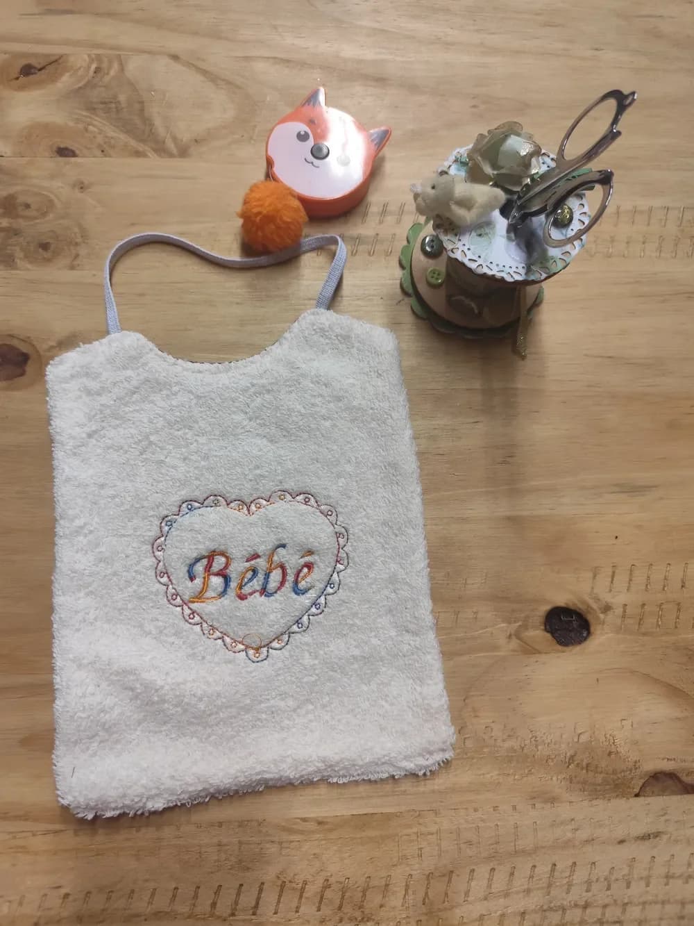 Bavoir en éponge écrue posé à plat sur une table en bois, brodé d'un grand cœur au contour festonné façon dentelle, avec au centre le mot « Bébé » en lettres calligraphiées, le tout en fil dégradé orange, bleu et rouge, avec un élastique gris clair pour passer autour du cou. Autour, un mètre ruban en forme de renard orange, un pompon orange et un pot à ciseaux décoré