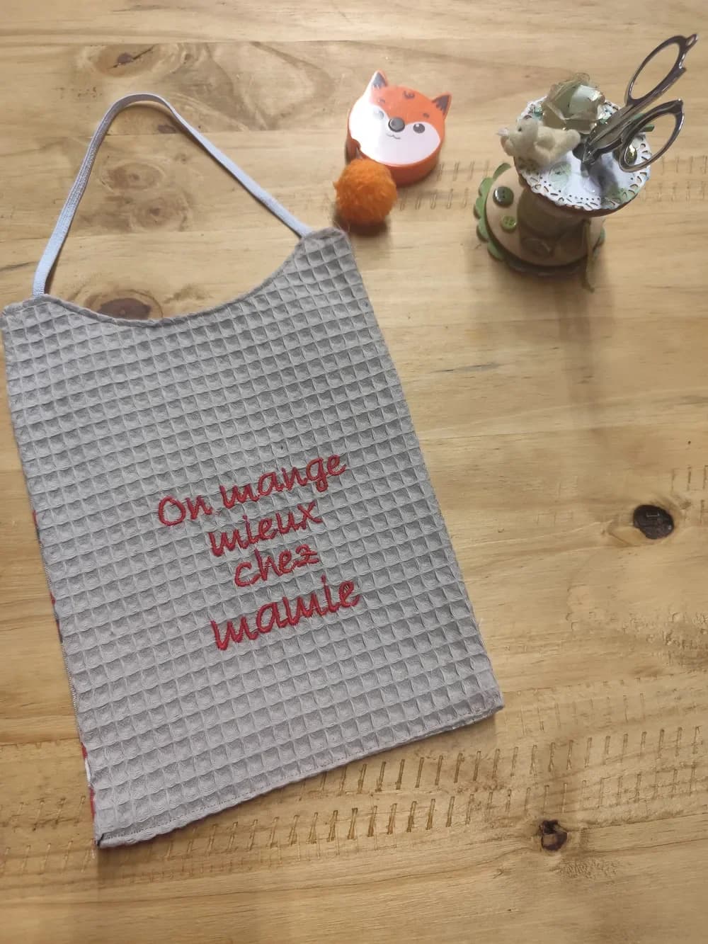 Bavoir en nid d'abeille gris clair posé à plat sur une table en bois, brodé en fil rouge sur quatre lignes du message « On mange mieux chez mamie » en lettres manuscrites, avec un élastique gris clair pour passer autour du cou. Autour, un mètre ruban en forme de renard orange, un pompon orange et un pot à ciseaux décoré