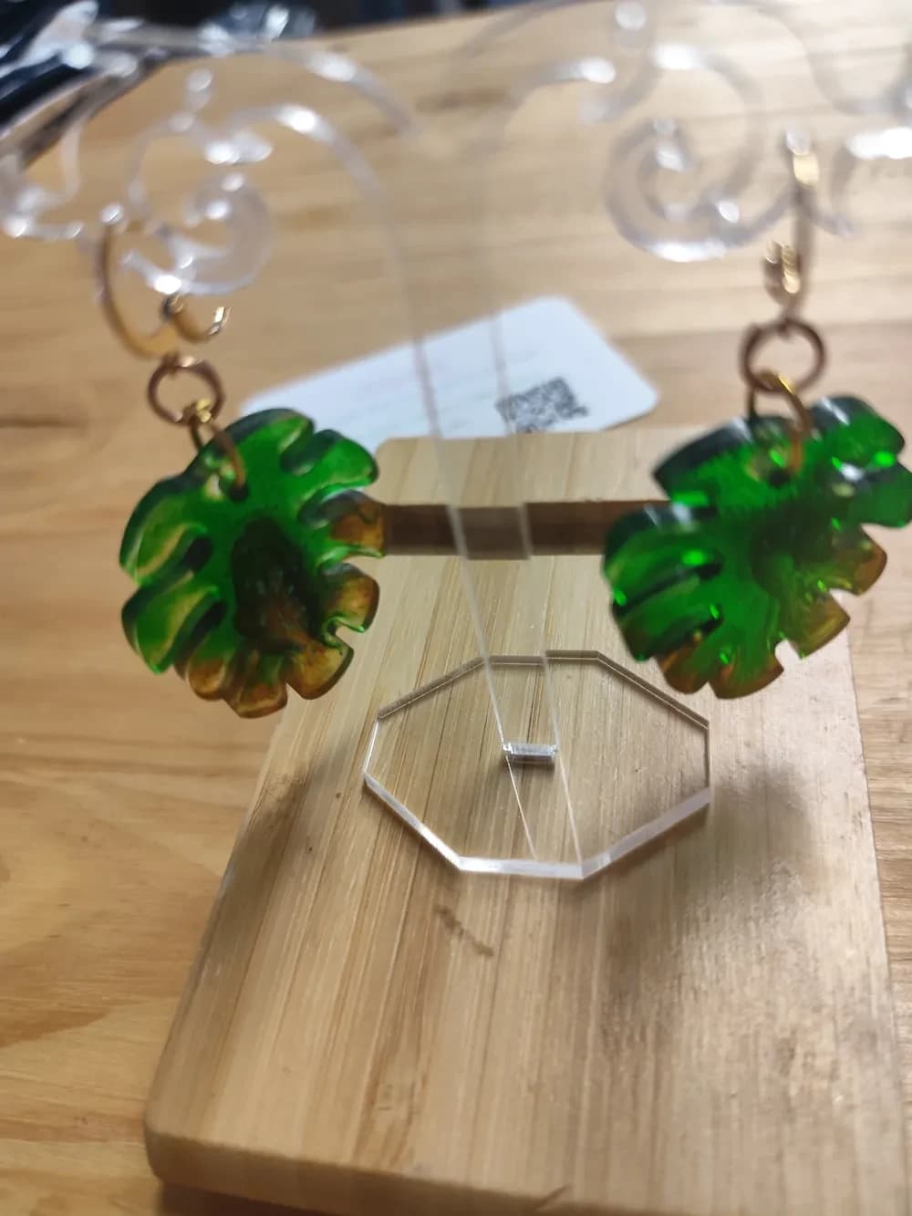 Paire de boucles d'oreilles en forme de feuille de monstera, une feuille tropicale arrondie aux bords profondément découpés en larges lobes. La résine translucide est d'un vert vif chiné, plus sombre au centre, et se teinte de nuances marron ambré sur le pourtour des lobes. Chaque feuille pend à un petit anneau et à un crochet doré à fermoir, accroché à un présentoir en plexiglas transparent. L'ensemble est posé sur une planche en bambou, devant une carte de visite floue.