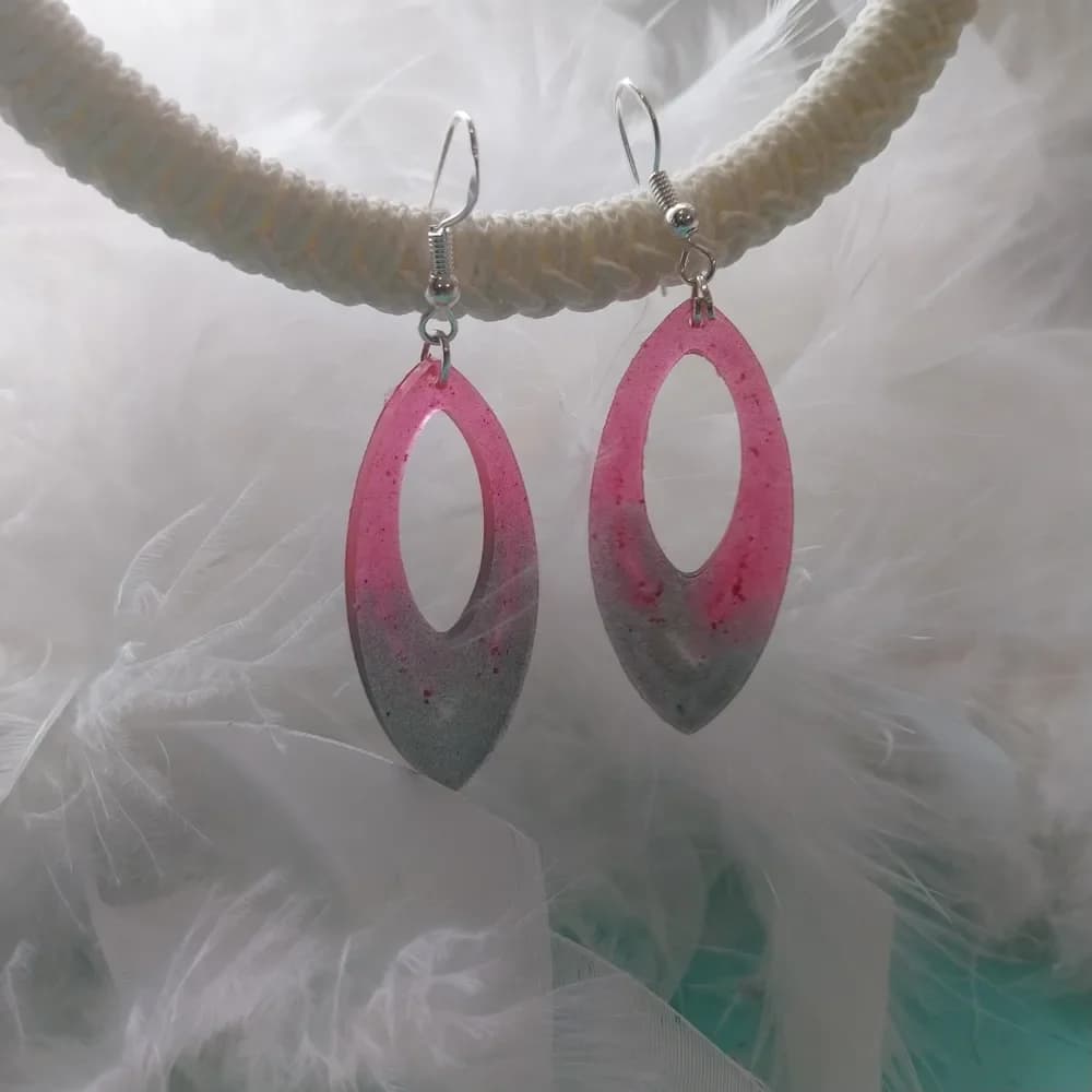 Paire de boucles d'oreilles en forme de marquise, un ovale allongé aux deux extrémités pointues, évidé au centre. La résine passe d'un rose framboise moucheté de petites taches rouge foncé, en haut, à un gris argenté pailleté vers la pointe du bas. Chaque boucle pend à un crochet argenté fixé par un petit anneau. Les deux boucles sont accrochées côte à côte à un anneau de corde tressée blanc cassé, au-dessus d'un nuage de plumes blanches, avec une touche de turquoise en bas de l'image.