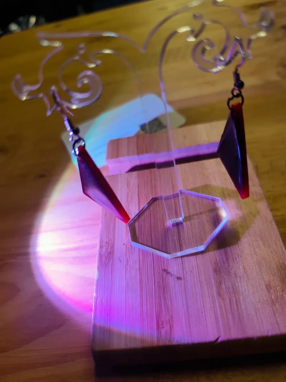Paire de boucles d'oreilles en résine en forme de triangle rectangle, suspendues par leur pointe à des crochets argentés, de chaque côté d'un présentoir en plexiglas transparent aux arabesques découpées. Le présentoir est posé sur une planche en bambou. La photo est prise sous un éclairage rose et violet qui donne aux triangles une teinte rouge sombre et prune : ce n'est pas leur vraie couleur, qui est orange et marron.