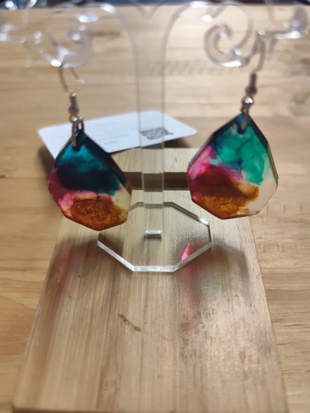 Paire de boucles d'oreilles en résine époxy transparente, de forme géométrique à facettes, suspendues par leurs crochets argentés à un présentoir en plexiglas transparent. Chaque boucle mêle en haut une tache vert canard profond, au milieu une touche de rose fuchsia et en bas un brun ambré aux reflets dorés, avec des zones laissées transparentes sur les bords. Le présentoir est posé sur une planche en bambou, devant une carte de visite floue.