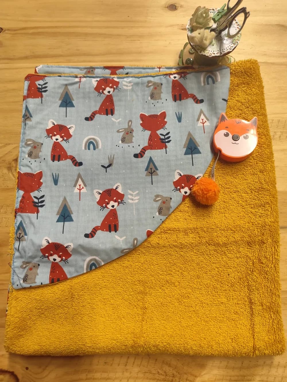 Cape de bain pour bébé en éponge jaune ocre, avec une capuche en coton bleu clair imprimé de petits renards rouge brun, de petits lapins et de petits arbres