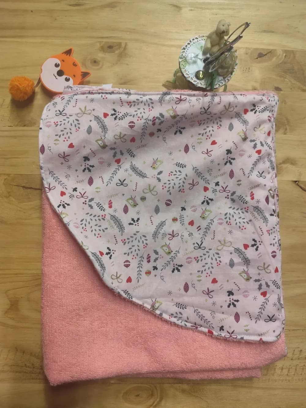 Cape de bain pour bébé en éponge rose saumoné, pliée et vue de face, avec sa capuche en coton rose pâle rabattue par-dessus, imprimée de fins motifs : feuillages gris, petits oiseaux, cœurs rouges, petits nœuds et brins de houx. Elle est posée sur une table en bois, à côté d'un mètre ruban en forme de renard orange.
