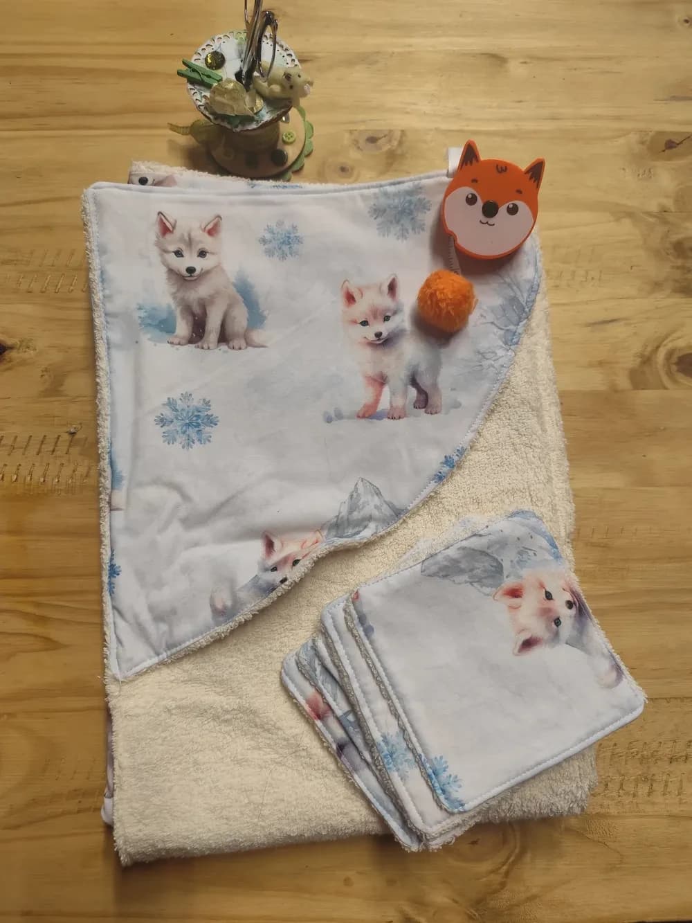 Cape de bain chien d'hiver dépliée à plat, montrant la capuche en coton imprimée de chiots blancs, de montagnes grises et de flocons de neige, doublée d'éponge crème, avec les six lingettes assorties disposées à sa droite et une décoration renard orange posée dessus