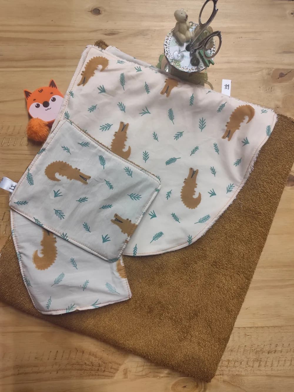 Tissu pour cape de bain bébé disposé à plat sur une table en bois : grand rectangle de coton écru à motif de crocodiles moutarde et de branches de sapin vertes, doublé de son revers en éponge de bambou ocre, accompagné de deux lingettes assorties au même imprimé et d'une décoration en forme de renard orange en arrière-plan