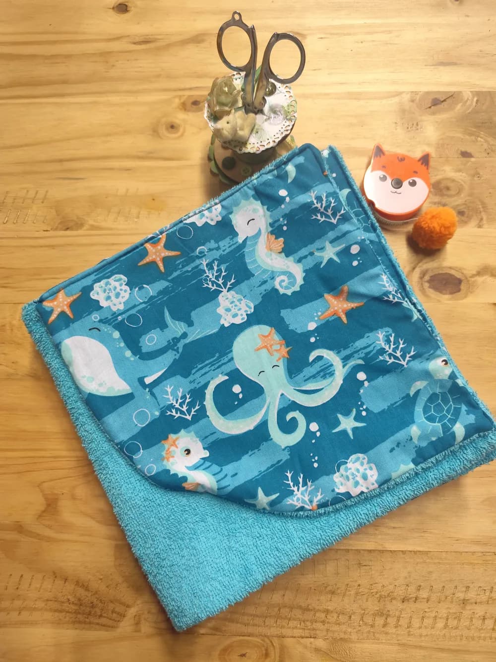 Cape de bain bébé pliée avec sa capuche pointue au premier plan, éponge bleu turquoise et face en coton imprimée d'un thème marin : hippocampes, pieuvres turquoise, étoiles de mer oranges, coraux blancs et bulles sur fond bleu à rayures, posée sur une table en bois avec une décoration en forme de renard orange