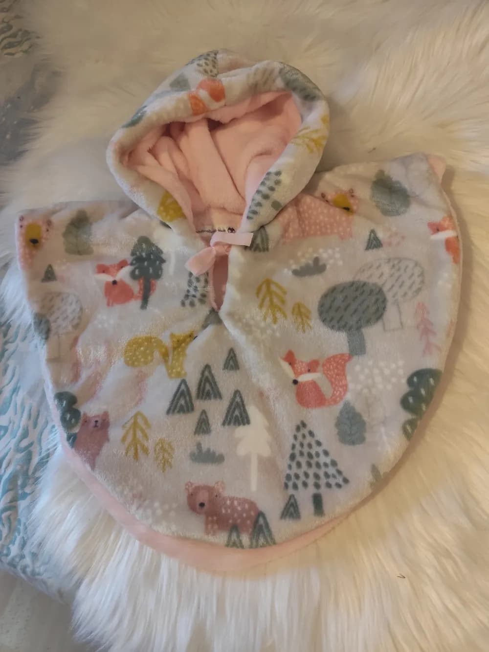 Cape bébé réversible Les animaux de la forêt CÉKIKAFÉ posée sur une fourrure blanche, capuche relevée, face blanc cassé imprimée de renards orangés, d'ours vieux rose, d'un écureuil moutarde, de sapins et d'arbres gris-vert et moutarde, intérieur rose très clair visible au revers de la capuche et sur le bord, petit lien rose noué au cou.