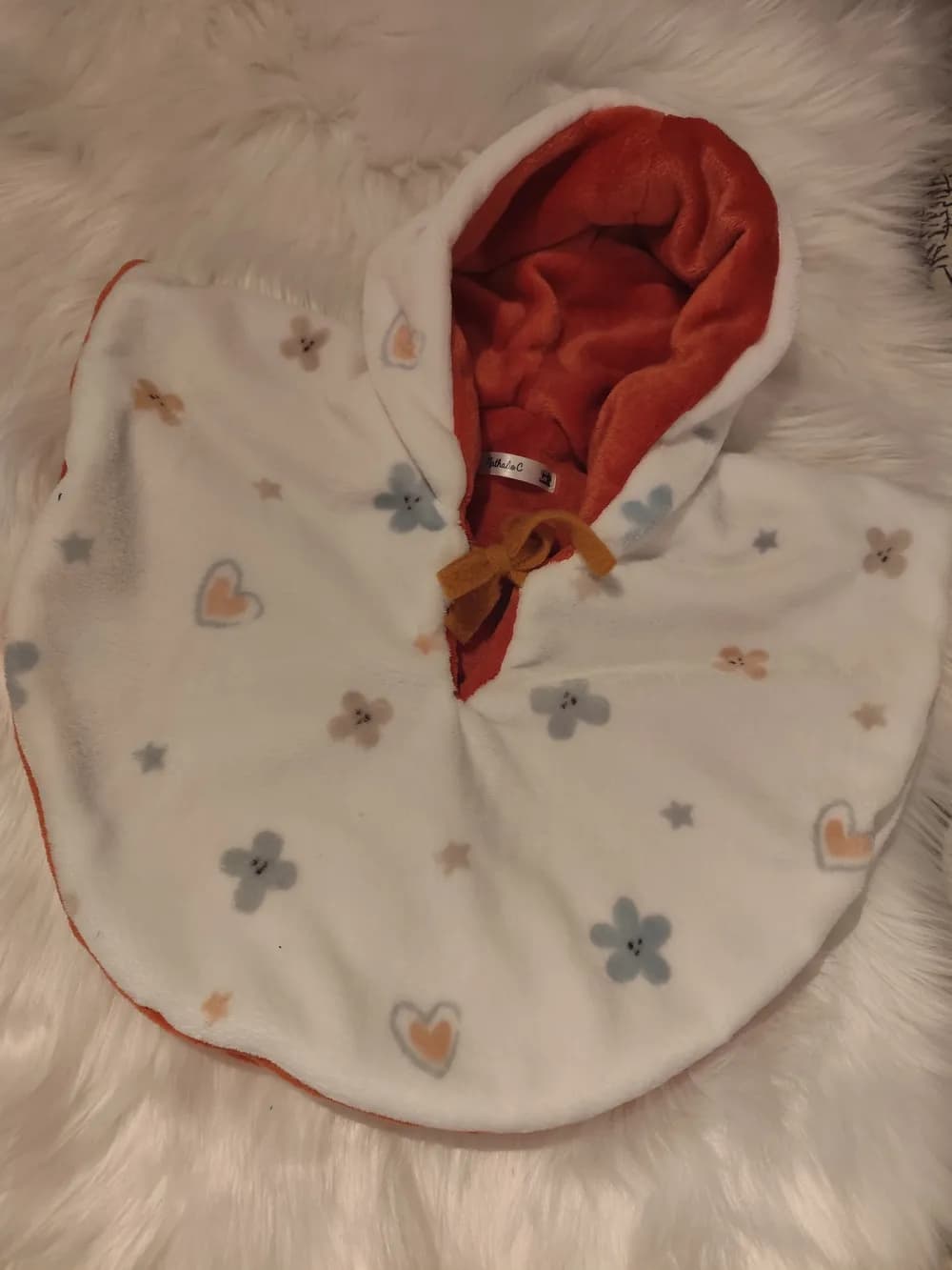 Cape bébé réversible Douceur fleurie CÉKIKAFÉ posée sur une fourrure blanche, capuche relevée, face blanche très douce semée de petites fleurs gris-bleu et beige à visage souriant, de cœurs beige cerclés de gris et de petites étoiles, capuche doublée de douillette terracotta et lien moutarde noué au cou.