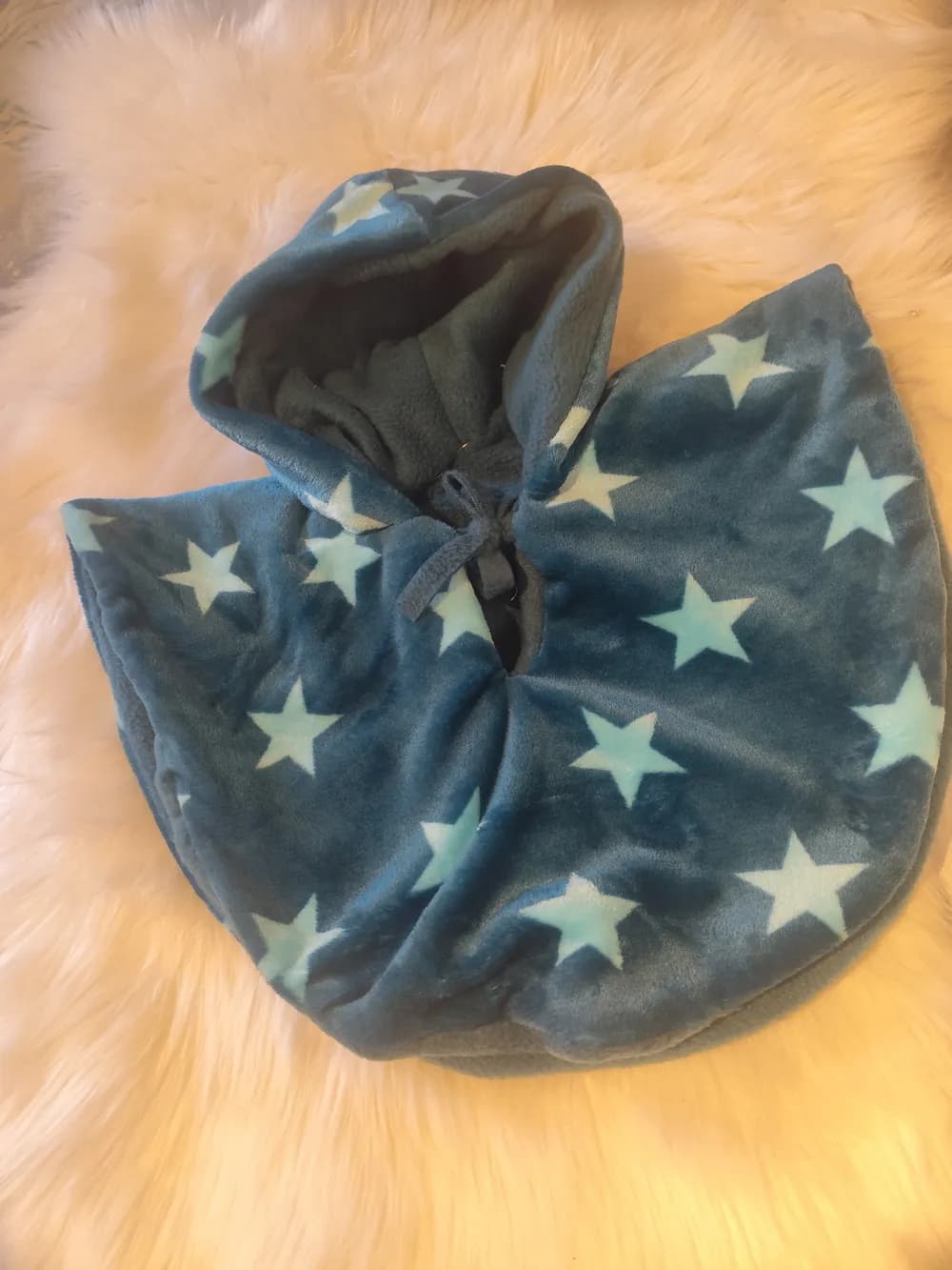 Cape bébé réversible CÉKIKAFÉ posée sur une fourrure blanche, capuche relevée, face bleue douce semée de grandes étoiles bleu clair, intérieur en polaire grise visible au revers de la capuche et sur le bord, petit lien noué au cou.