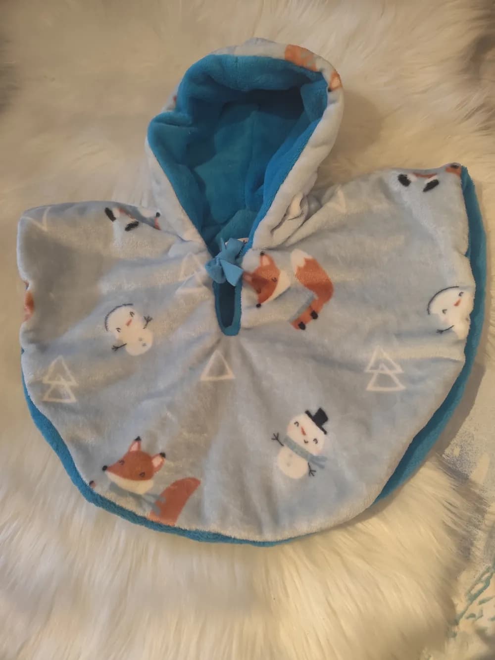 Cape bébé réversible L'hiver arrive CÉKIKAFÉ posée sur une fourrure blanche, capuche relevée, face en polaire gris clair bleuté imprimée de petits renards roux en écharpe, de bonhommes de neige et de triangles blancs, capuche et bord doublés de polaire bleu turquoise, petit nœud turquoise au cou.