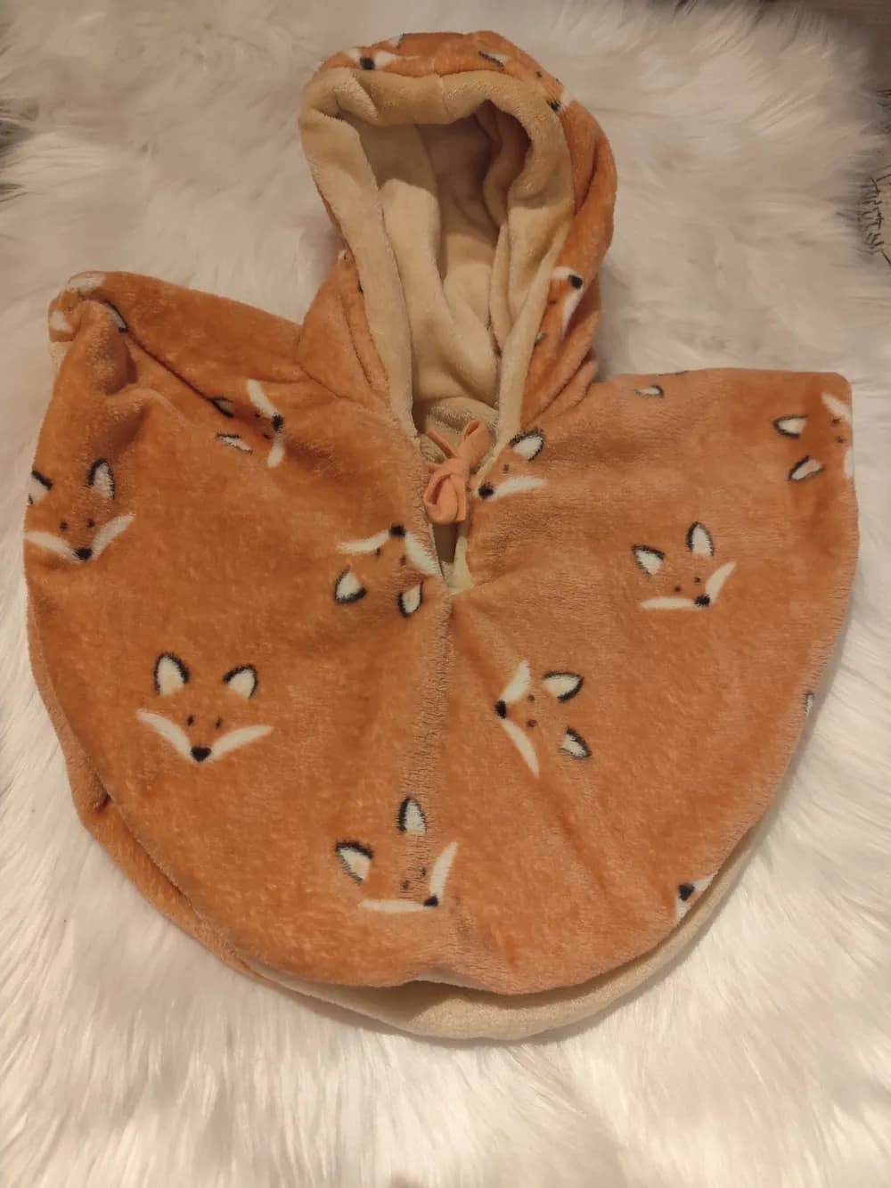 Cape bébé réversible La Tête du Renard posée sur une fourrure blanche, capuche relevée, face en douillette ocre orangé ornée de têtes de renard blanches aux oreilles noires, capuche doublée de douillette beige et petit lien orangé noué au cou.
