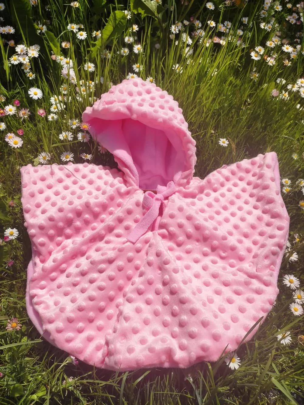 Cape bébé réversible CÉKIKAFÉ en Minky rose clair à petits pois en relief, capuche relevée, posée dans l'herbe au milieu des pâquerettes, intérieur en polaire rose visible dans la capuche.