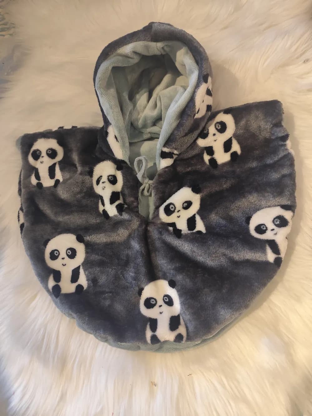 Cape bébé réversible Le panda tout doux CÉKIKAFÉ posée sur une fourrure blanche, capuche relevée, face en douillette gris anthracite épaisse imprimée de pandas noir et blanc assis et souriants, intérieur en polaire douillette vert d'eau pâle visible au revers de la capuche, lien vert d'eau noué au cou.