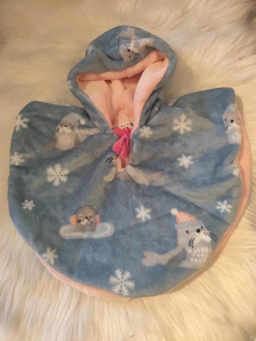 Cape bébé réversible Petit phoque des neiges CÉKIKAFÉ posée sur une fourrure blanche, capuche relevée, face bleu-gris semée de grands flocons blancs et de phoques gris, l'un coiffé d'un bonnet rose à pompon, un autre allongé sur un nuage avec un cache-oreilles orange, capuche doublée de rose pâle et nœud rose fuchsia au cou.