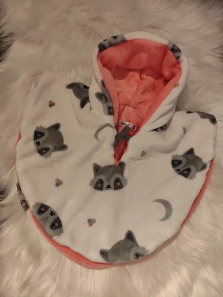Cape bébé réversible Petit raton laveur CÉKIKAFÉ posée sur une fourrure blanche, capuche relevée, face blanche semée de têtes de raton laveur grises au masque noir, de croissants de lune gris et de petits points, capuche doublée de douillette pêche saumon et lien gris noué au cou.