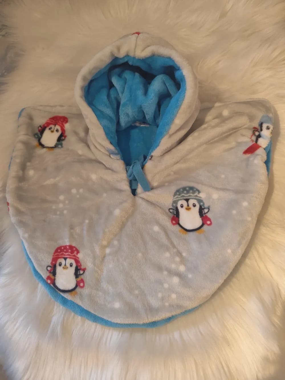 Cape bébé réversible Pingouins des neiges CÉKIKAFÉ posée sur une fourrure blanche, capuche relevée, face gris clair semée de flocons blancs et de pingouins coiffés de bonnets rouges ou gris-bleu avec moufles rouges, capuche doublée de polaire douillette bleu turquoise et lien turquoise noué au cou.