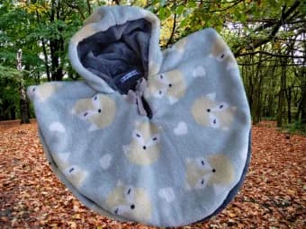 Cape bébé réversible présentée capuche relevée, en forme de petite cape arrondie. Face extérieure en polaire douillette gris clair légèrement bleuté, ornée de jolies têtes de renards beige et de petits cœurs blancs. L'intérieur de la capuche laisse voir la polaire gris plus foncé et soyeuse de l'autre face, avec un petit lien noué au cou. La cape se détache sur un décor de forêt d'automne aux feuilles mortes orangées