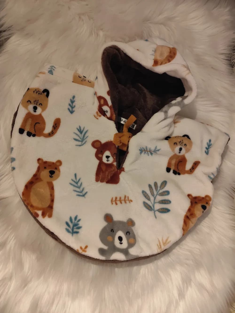 Cape bébé réversible La ronde des ours CÉKIKAFÉ posée sur une fourrure blanche, capuche relevée, face blanche imprimée d'ours roux, d'ours caramel tachetés, d'un ours gris et de petits félins tachetés assis, parmi des branches bleu canard et ocre, intérieur marron chocolat visible au revers de la capuche et lien moutarde noué au cou.