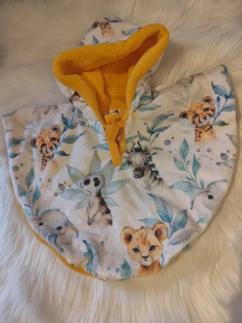 Cape bébé réversible Safari CÉKIKAFÉ posée sur une fourrure blanche, capuche relevée, face en Minky blanc imprimé à l'aquarelle d'un lionceau, de guépards, d'un zèbre, d'un suricate et d'un éléphanteau parmi des feuillages bleu-vert, capuche doublée de douillette jaune soleil et lien jaune noué au cou.