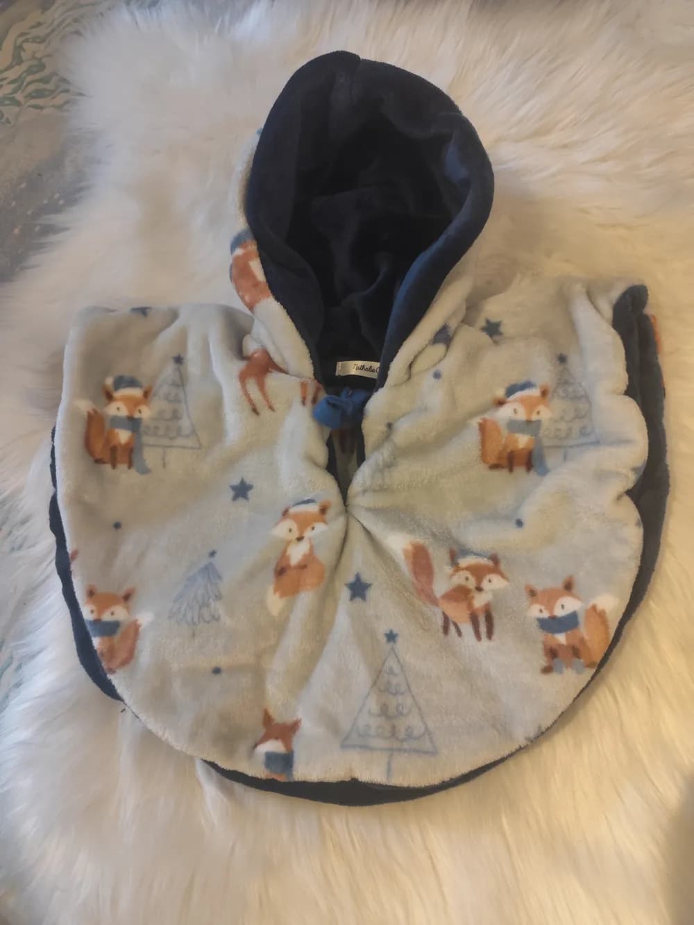 Cape bébé réversible La valse des renards CÉKIKAFÉ posée sur une fourrure blanche, capuche relevée, face gris clair bleuté imprimée de petits renards roux coiffés de bonnets bleus avec écharpes, de sapins dessinés en bleu et d'étoiles bleues, capuche et bord doublés de polaire douillette bleu marine, lien bleu noué au cou.