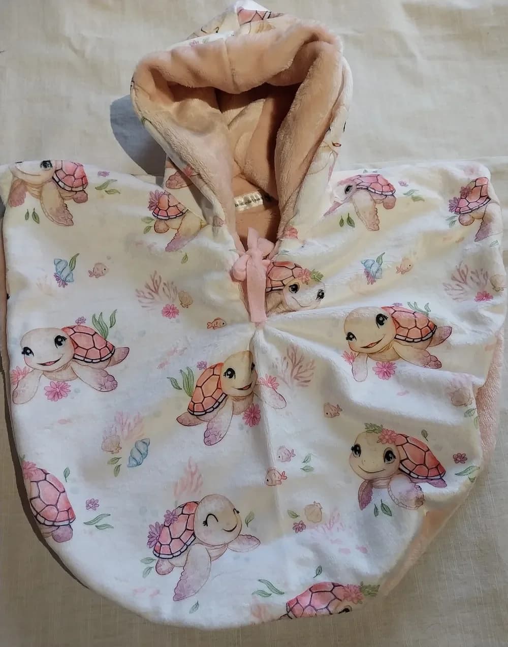 Cape réversible pour bébé posée à plat sur un drap crème, capuche relevée, face en minky blanc imprimé de tortues roses souriantes entourées de fleurs, de coraux et de coquillages, doublure en polaire rose clair et petit lien rose noué au cou