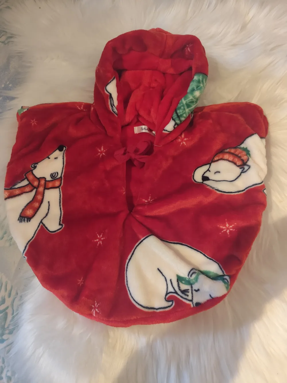 Cape réversible bébé douillette rouge ours polaires – CÉKIKAFÉ. La cape posée sur une fourrure blanche, capuche relevée, côté douillette rouge imprimée d'ours polaires blancs en écharpe et bonnet de Noël et de petits flocons ; la capuche est doublée de douillette rouge unie et un lien rouge est noué au cou.