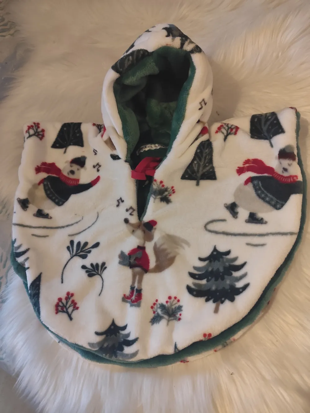 Cape polaire réversible bébé ours patineur, couverture de cosy – CÉKIKAFÉ. La cape posée sur une fourrure blanche, capuche relevée, face polaire blanche imprimée d'ours et de renards patineurs en écharpe rouge, de sapins, de baies rouges et de notes de musique ; la capuche et le bord laissent voir une doublure verte unie, et un lien rouge est noué au cou.