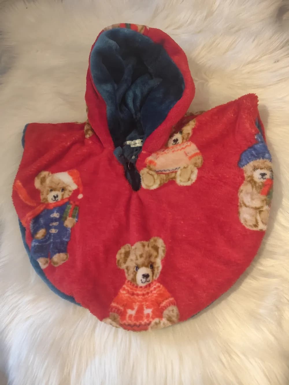 Cape réversible bébé The Bear polaire rouge oursons intérieur bleu canard – CÉKIKAFÉ. La cape posée sur une fourrure blanche, capuche relevée, côté polaire rouge imprimée d'oursons en pull de Noël, en bonnet rouge et en manteau bleu ; la capuche laisse voir la douillette bleu canard de l'intérieur.
