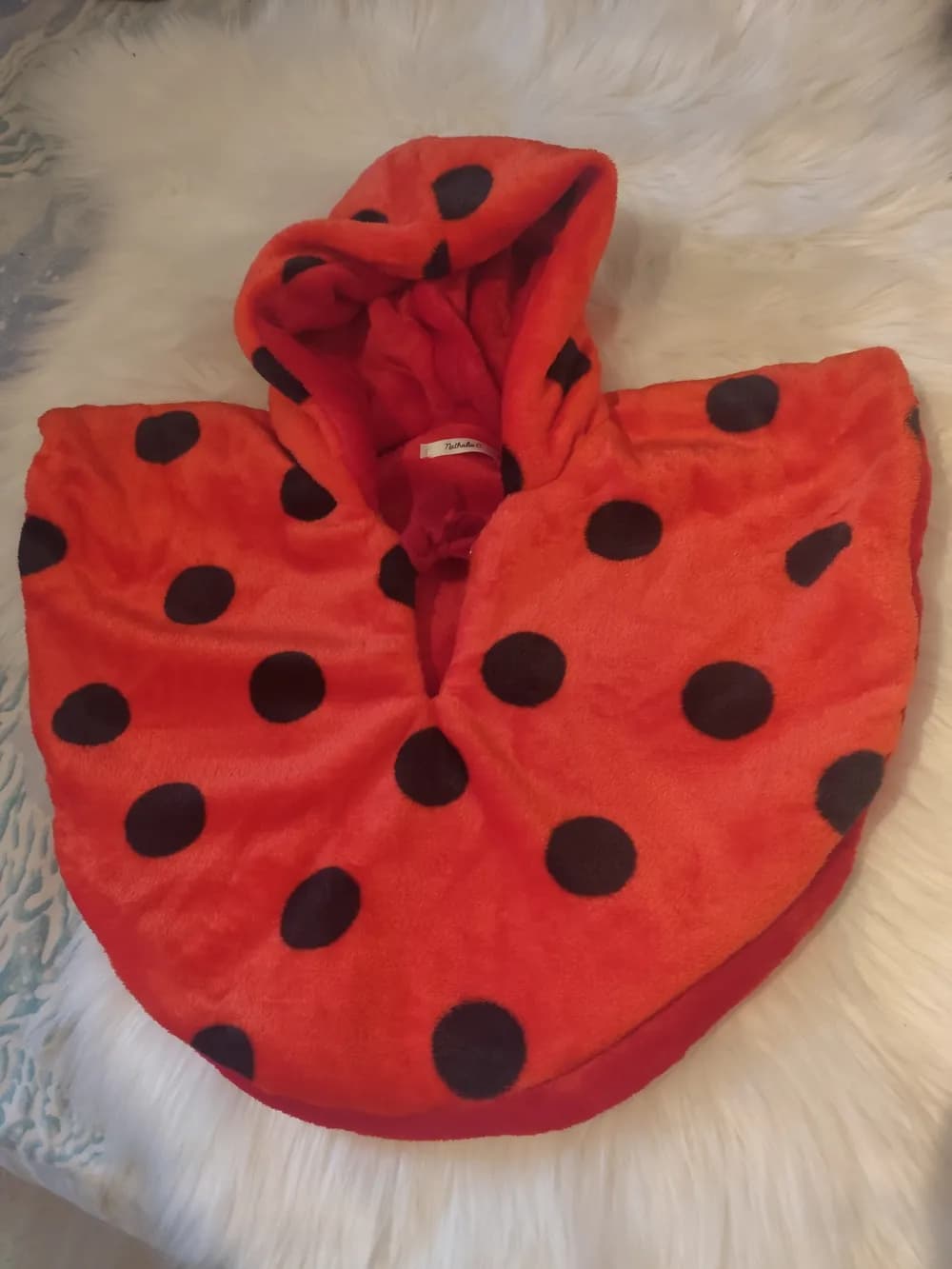 Cape réversible Petit Chaperon Rouge CÉKIKAFÉ posée sur une fourrure blanche, capuche relevée, face rouge vif semée de gros pois noirs, intérieur en polaire douillette rouge visible au bord, petite étiquette cousue sous la capuche.