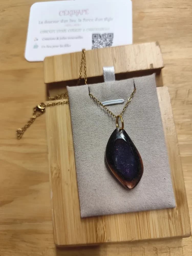 Collier présenté à la lumière naturelle sur un présentoir en suédine grise, posé sur une planche en bambou, avec une carte de visite CÉKIKAFÉ en arrière-plan. Le pendentif, en forme de galet ovale légèrement pointu vers le bas, est en résine brun très foncé, translucide et cuivrée sur les bords. En son centre, un creux ovale est rempli d'une résine bleu violacé parsemée de fines paillettes scintillantes, comme un petit lac. Il pend par un anneau doré à une fine chaînette dorée à maillons, dont le fermoir mousqueton est visible à gauche.