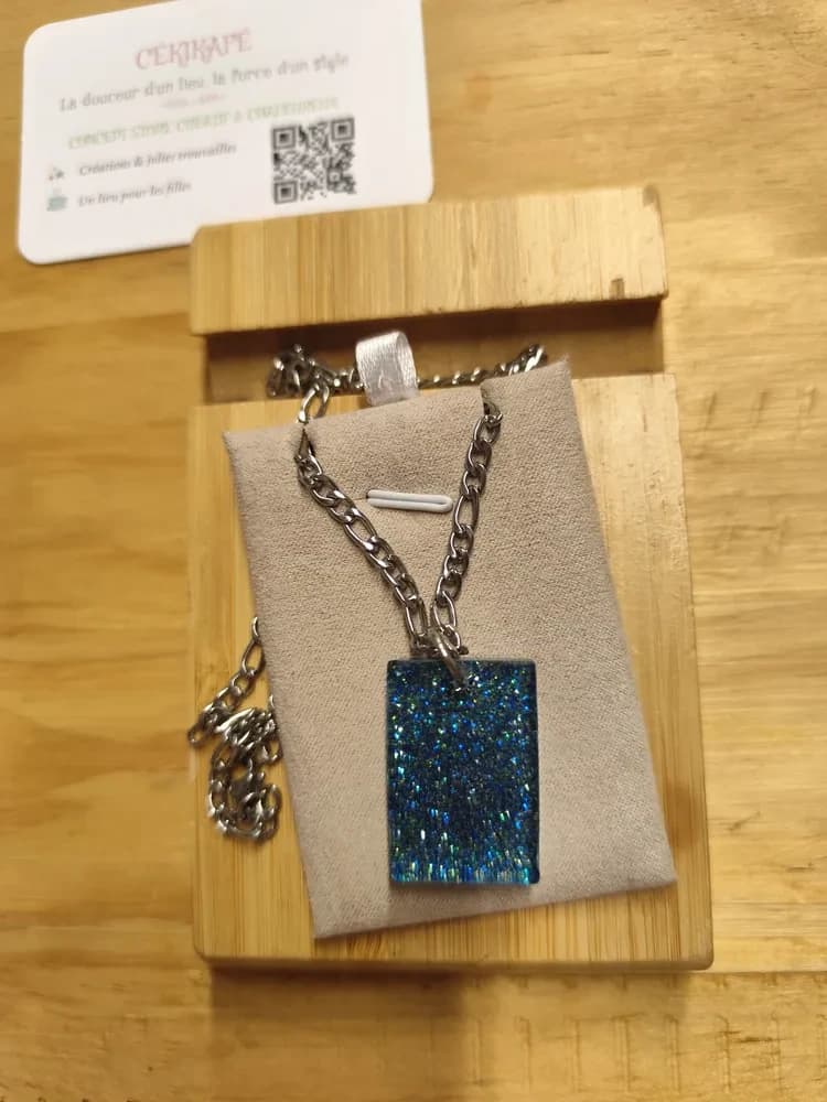 Collier présenté à la lumière naturelle sur un présentoir en suédine grise, posé sur une planche en bambou, avec une carte de visite CÉKIKAFÉ en arrière-plan. Le pendentif est un rectangle vertical en forme de lingot, en résine transparente remplie de paillettes bleu foncé, bleu clair et vert d'eau, dispersées et scintillantes. Il pend par un anneau à une chaîne argentée assez épaisse, à maillons plats alternant courts et longs, dont le fermoir mousqueton repose sur la gauche de la planche.