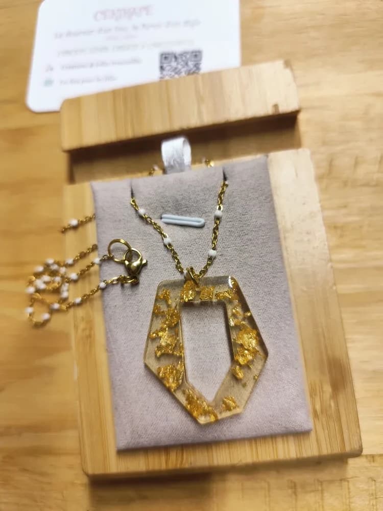 Collier présenté à la lumière naturelle sur un présentoir en suédine grise, posé sur une planche en bambou, avec une carte de visite CÉKIKAFÉ en arrière-plan. Le pendentif est un losange allongé, pointe vers le bas, aux côtés facettés comme une petite couronne, ajouré au centre. Sa résine transparente est parsemée de nombreux éclats de papier doré. Il pend par un anneau doré à une fine chaînette dorée ponctuée de petites perles blanches. À gauche, le large fermoir mousqueton doré et son anneau reposent sur le présentoir.