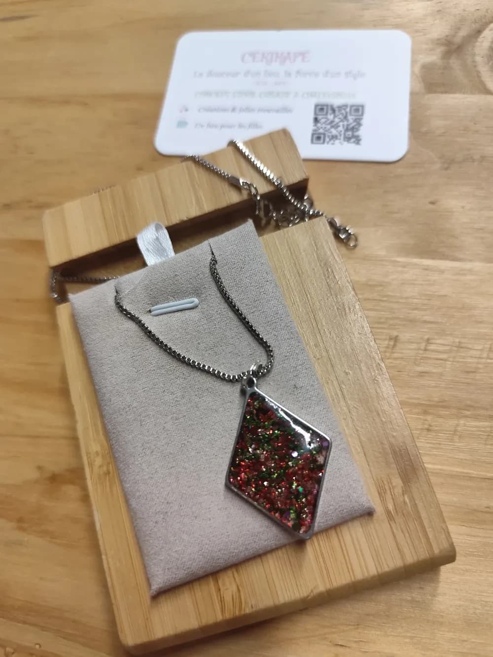 Collier présenté à la lumière naturelle sur un présentoir en suédine grise, posé sur une planche en bambou, avec une carte de visite blanche CÉKIKAFÉ en arrière-plan. Le pendentif en losange, pointe vers le bas, est suspendu par un petit anneau à une chaînette argentée à mailles carrées. Son cadre en métal argenté brillant est rempli de résine où se mêlent une majorité de paillettes rouges, des éclats verts et quelques touches dorées. Le fermoir et la rallonge de la chaînette dépassent en haut à droite.