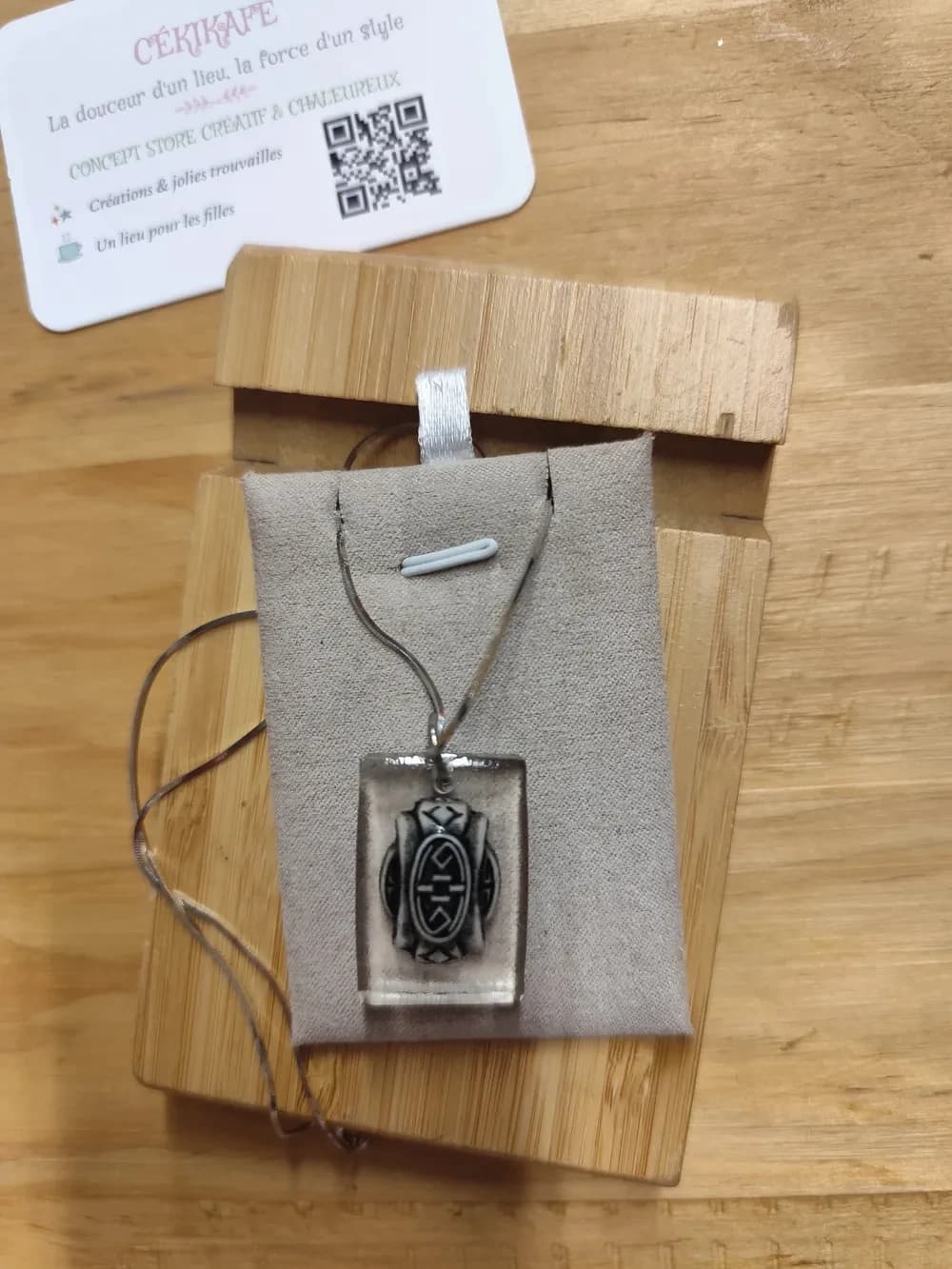 Collier photographié de près à la lumière naturelle, sur un présentoir en suédine grise posé sur une planche en bambou, sur une table en bois clair. Le pendentif rectangulaire aux coins arrondis est en résine transparente comme du verre. La perle enfermée à l'intérieur est ivoire veinée de gris, sculptée en relief : un ovale central noir avec des lignes et des crochets, entouré de chevrons en haut et en bas et de deux disques sur les côtés. La chaînette fine argentée passe dans un petit anneau au sommet et retombe en boucles sur la gauche. Carte de visite CÉKIKAFÉ en haut à gauche.