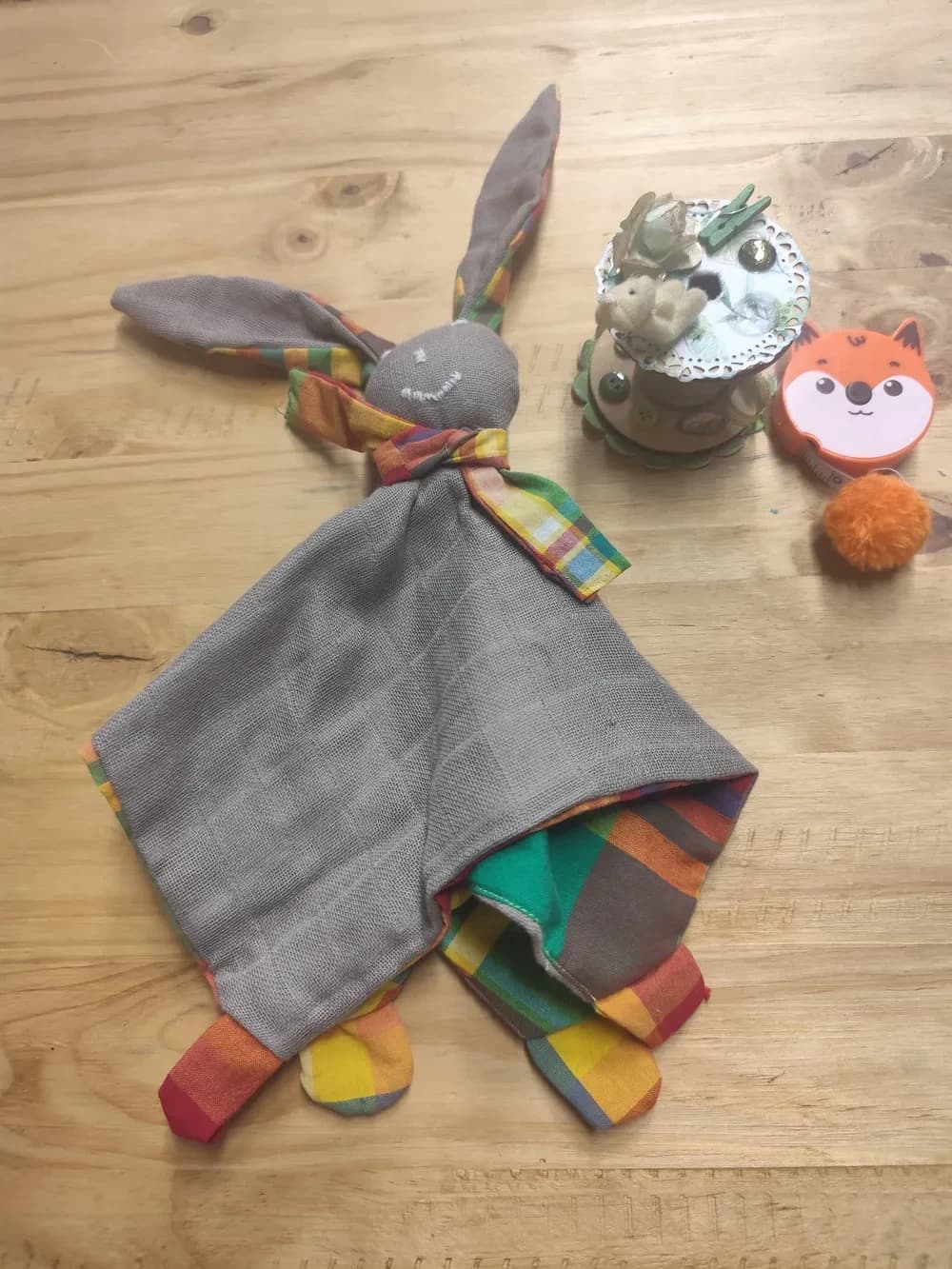 Doudou carré fait main CÉKIKAFÉ en gaze de coton taupe, avec en haut une tête ronde aux yeux et à la bouche brodés à la main en fil blanc, deux grandes oreilles de lapin dressées doublées de madras, une petite écharpe cousue en madras à carreaux jaunes, verts, rouges et bleus, et une patte en madras dans chaque coin. Le bas du carré est replié et laisse voir le madras coloré du dessous. Posé sur une table en bois clair.