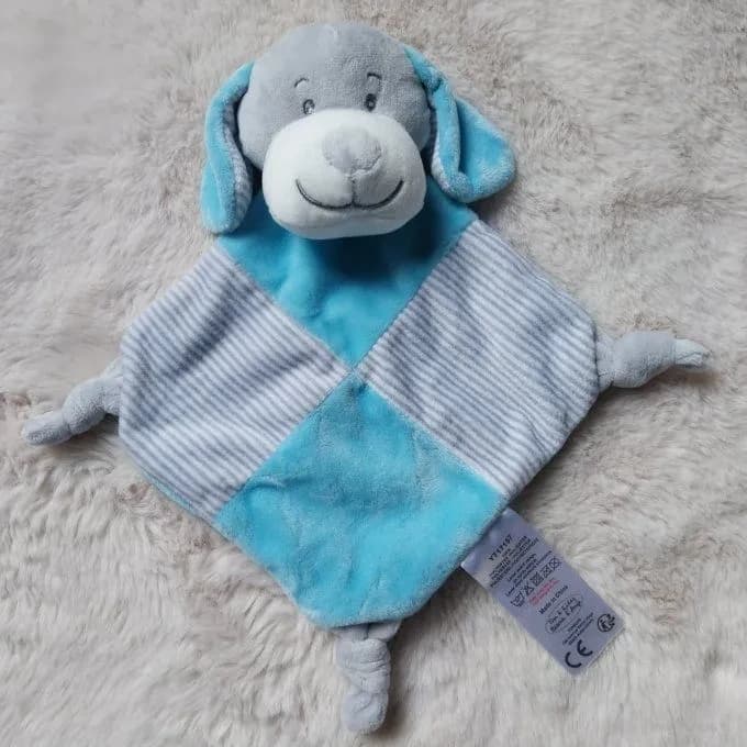Doudou chien version bleue, à personnaliser avec un prénom brodé, posé à plat sur une fourrure beige : une tête de chien en peluche grise au museau blanc, au nez gris et au sourire brodé, avec deux oreilles tombantes bleu turquoise. Dessous, un carré posé en losange qui alterne velours bleu turquoise et tissu rayé gris et blanc, avec un petit nœud gris à chaque coin. Une étiquette blanche avec le logo CE est cousue sur le côté.