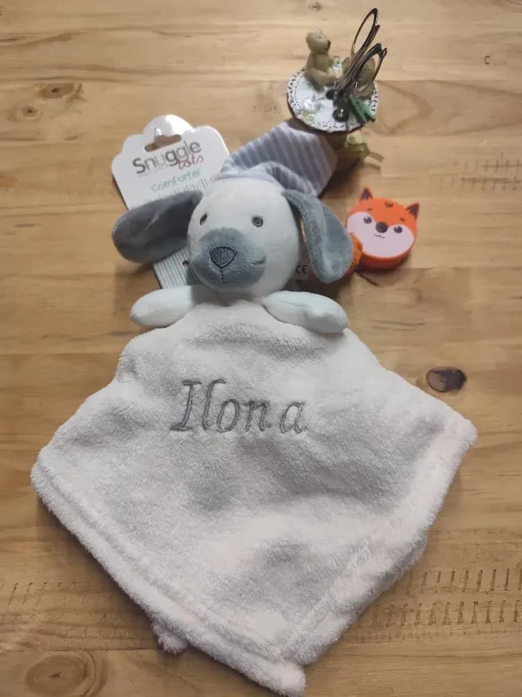 Doudou Snuggle Tots en douillette crème, avec une tête de chien blanc cassé, des oreilles et un museau gris, des yeux et un nez brodés, deux petits bras et un bonnet de nuit rayé gris-bleu et blanc. Le prénom « Ilona » est brodé en fil gris sur le carré. L'étiquette cartonnée Snuggle Tots « Comforter » est encore attachée. Posé sur une table en bois, à côté d'un petit renard orange décoratif.