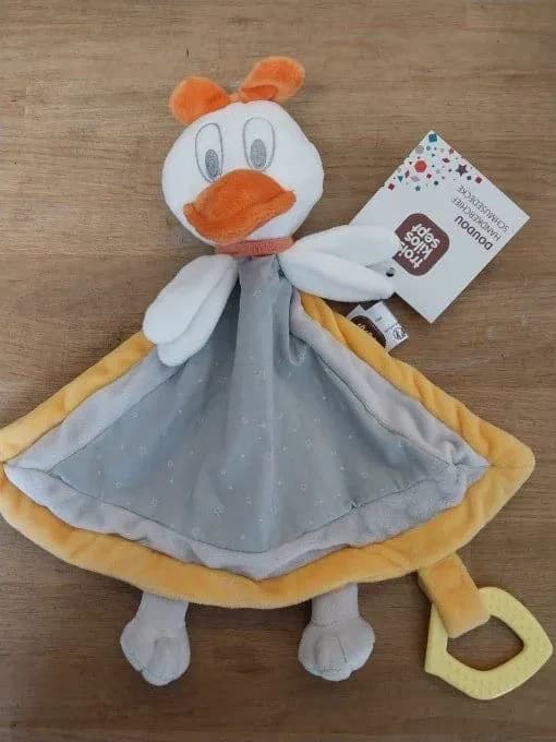 Doudou Daisy de la marque Trois Kilos Sept, avec de nombreuses prises pour les petites mains et un anneau de dentition en bas. Daisy est une petite cane : tête blanche en peluche, grands yeux brodés en gris, bec orange, nœud orange sur la tête et collier corail, deux ailes blanches. Son corps est un carré de velours gris clair parsemé de petites étoiles blanches, bordé de jaune moutarde, avec deux pattes grises en bas et un anneau de dentition jaune pâle accroché à un ruban jaune. L'étiquette Trois Kilos Sept est encore attachée. Posée sur une table en bois.