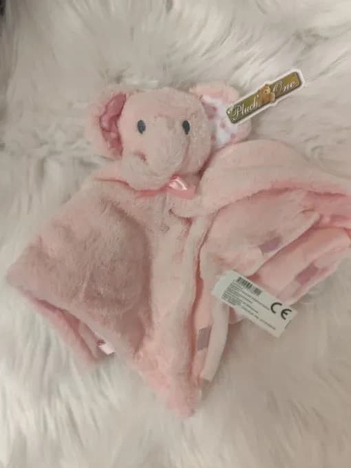 Doudou hochet éléphant rose de la marque Pluch'One : une tête d'éléphant cousue au centre d'un carré tout doux, avec des oreilles dont l'intérieur est d'une autre couleur. Peluche rose pâle à poils doux, petits yeux noirs, trompe, intérieur des oreilles rose vif à pois blancs et petit nœud de satin rose sous la tête ; le carré est replié et bordé de petites boucles de ruban rose. L'étiquette Pluch'One et l'étiquette CE sont encore attachées. Posé sur une fourrure blanche.