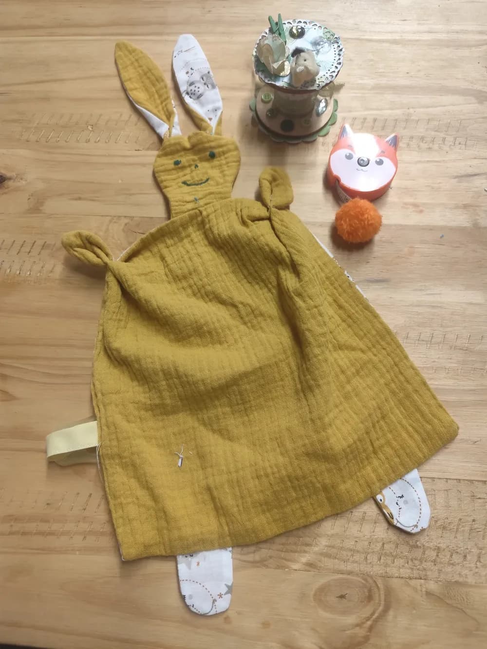 Doudou lapin plat fait main CÉKIKAFÉ en gaze de coton ocre doré, avec une tête plate aux deux yeux et au sourire brodés en fil vert, deux grandes oreilles ocre doublées de coton blanc imprimé, deux petits bras noués en haut, deux petites pattes en coton blanc imprimé en bas et un ruban jaune pâle sur le côté. Posé sur une table en bois clair, à côté d'une bobine décorée et d'un mètre ruban en forme de renard orange.