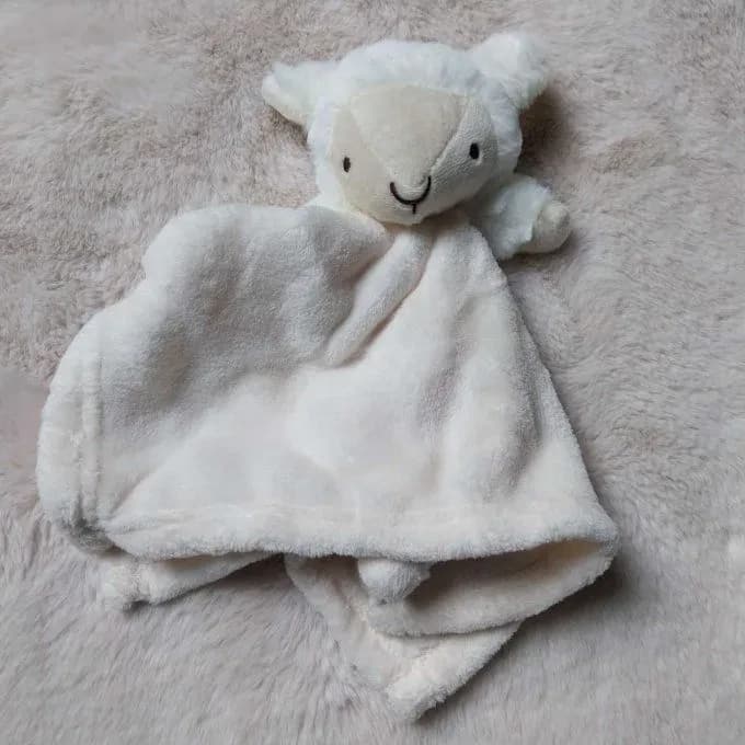 Doudou petit mouton Snuggle Tots posé sur une fourrure synthétique beige rosé, vu de face. En haut, une tête de mouton toute ronde en peluche bouclée blanc crème, avec deux petites oreilles, un museau beige clair en forme de triangle, deux yeux noirs et un sourire brodés en fil noir, et deux petites pattes de chaque côté. En dessous, le carré de douillette crème très doux retombe en plis. Aucun prénom n'est brodé.