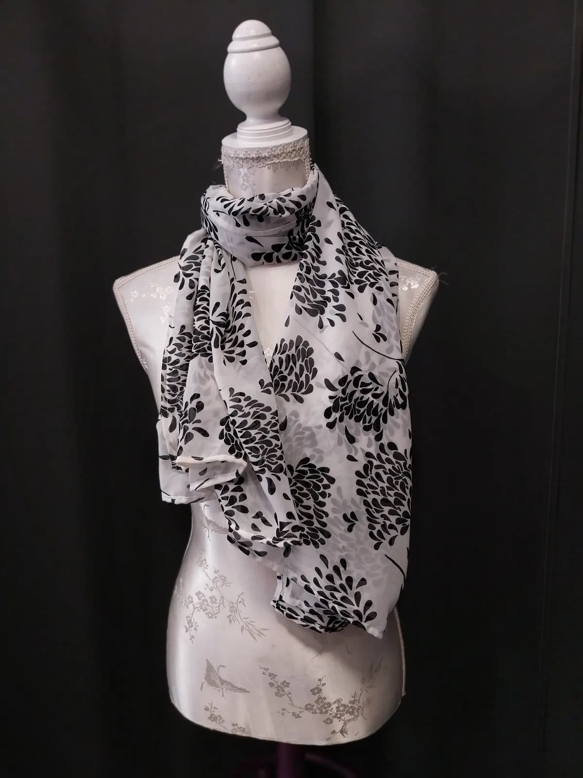 Foulard blanc à motifs floraux noir et gris façon fleurs stylisées, drapé et noué autour du cou d'un buste mannequin, pans retombant jusqu'à la taille