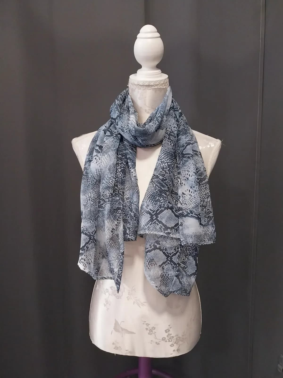 Foulard fin et souple à effet peau de serpent, dans un dégradé de gris, de noir et de blanc, enroulé une fois autour du cou d'un buste de couture blanc, ses deux pans tombant librement devant, sur un fond gris anthracite