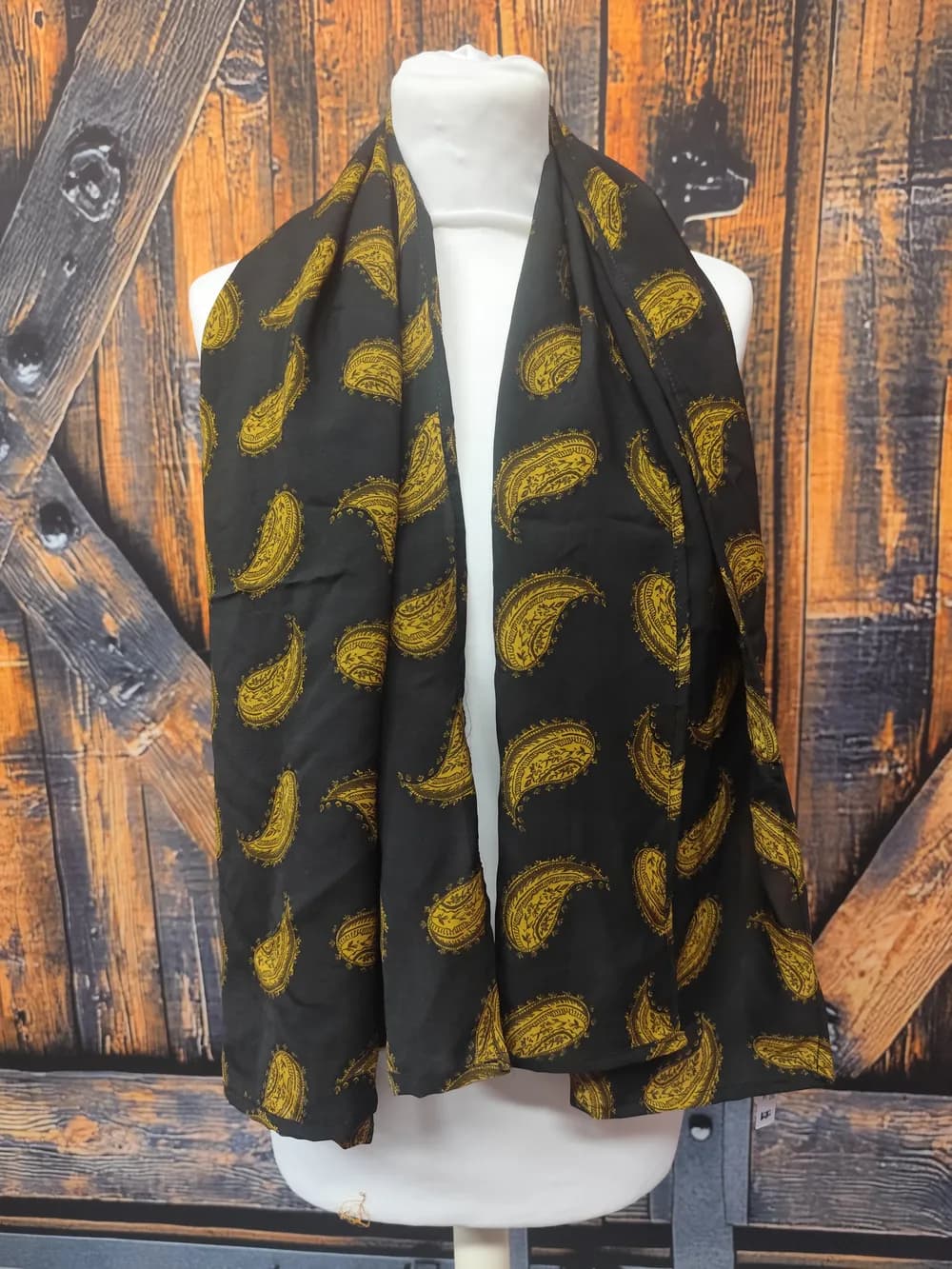 Foulard noir à motif façon plumes dorées, drapé et noué autour du cou d'un buste mannequin, pans longs retombant jusqu'à la taille, devant un fond de planches de bois brutes orangées